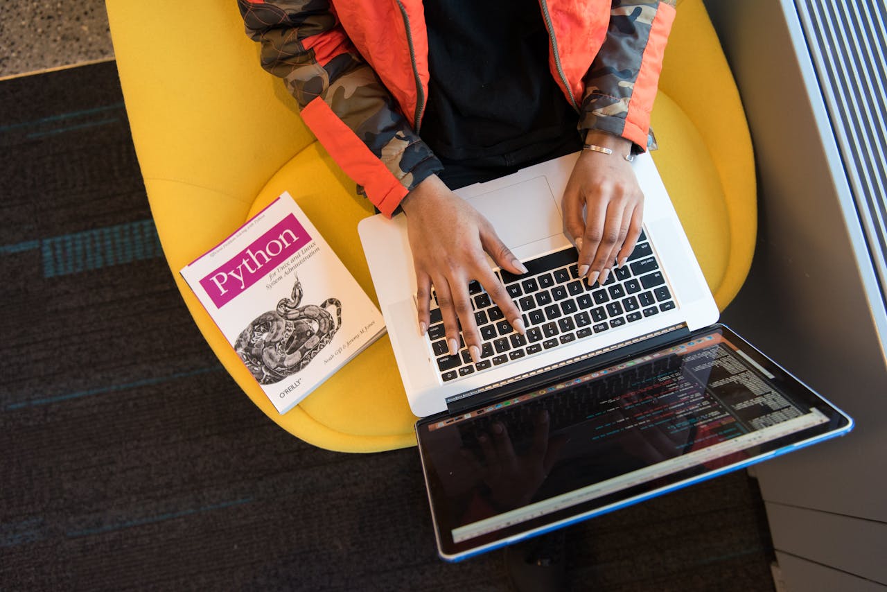 creative A developer typing code on a laptop with a Python book beside in an office.